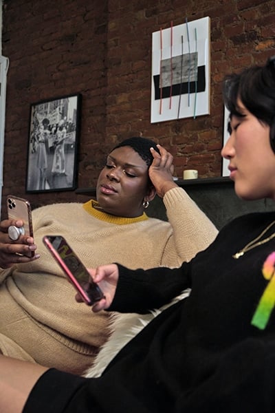 Una mujer trans negra no binaria y una persona asiática no binaria utilizando sus teléfonos móviles.