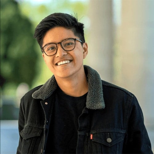 Alex del Rosario, une joven filipine trans no binarie, sonríe a la cámara. Pelo corto y oscuro, gafas, camisa negra y chaqueta vaquera con cuello gris. Fondo verde y claro suavemente difuminado.