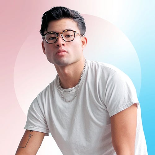 Chella Man, a transgender, Jewish, Chinese, and deaf actor/artist/producer, looks calmly at the camera. Short dark hair, round glasses, a silver chain, white t-shirt. Tattoo on forearm. Soft pink-to-blue gradient background with a central white circle.