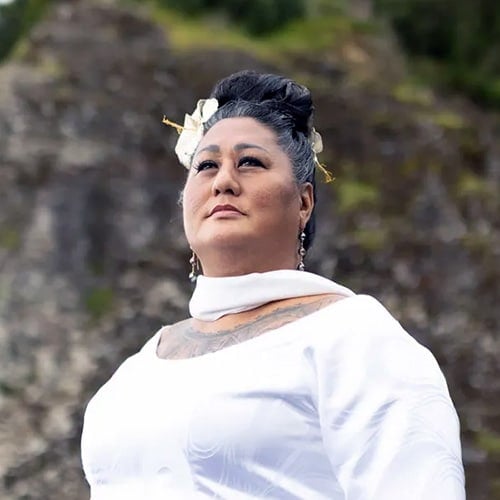 Kumu Hina mira regiamente hacia arriba. Su cabello oscuro está peinado con flores blancas y doradas. Lleva un vestido blanco que deja ver un tatuaje tradicional. Fondo rocoso y verde suavemente difuminado.